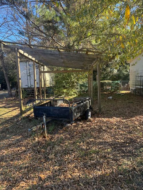 A dark utility trailer sits parked under a wooden, open-air shelter in a yard covered with fallen autumn leaves.