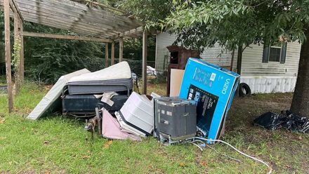 A pile of discarded household items, including a large blue TV box and a mattress, sits in a grassy yard under a canopy.