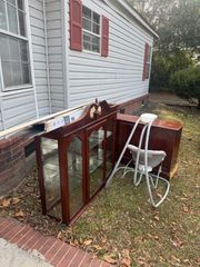 A wooden display cabinet and a baby swing sit on a grassy yard next to the brick foundation of a light-sided house.