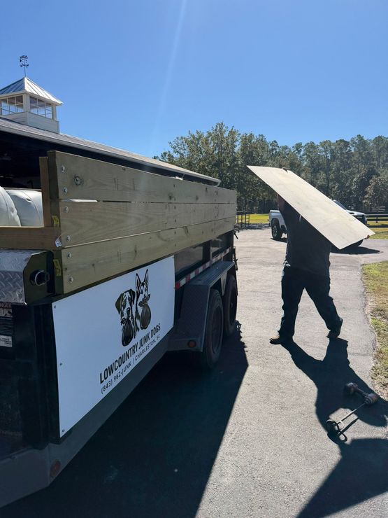 A person in dark clothes carries a large wooden board toward the back of a dump trailer with a company logo on the side.