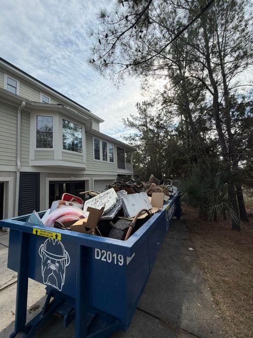 A large blue dumpster filled with household trash sits on a driveway next to a light-colored multi-story house.