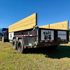 A dark truck pulls a dual-axle dump trailer with wood sideboards, featuring dog-themed branding, on a grassy field.