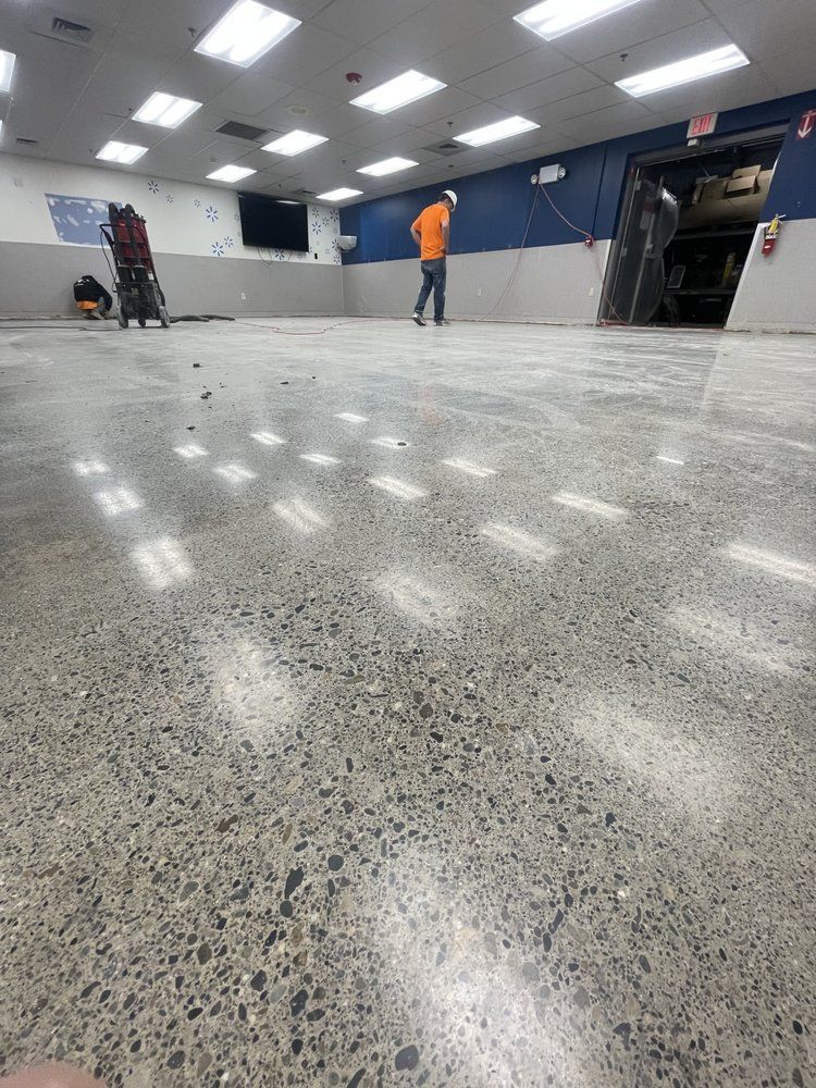 Large empty indoor room with gray speckled floor, bright ceiling lights, and two people near the back wall