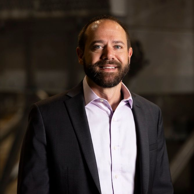 Man in blazer and pink shirt, smiling with a beard, indoors.