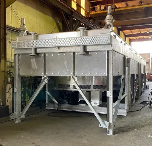Large stainless-steel industrial machine on a factory floor, with angled support legs and overhead piping