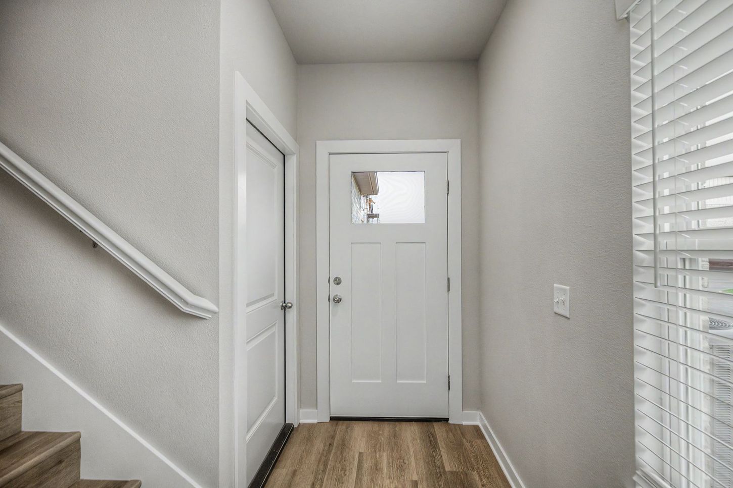 Narrow hallway with stairs, white front door, and a window with blinds on the right