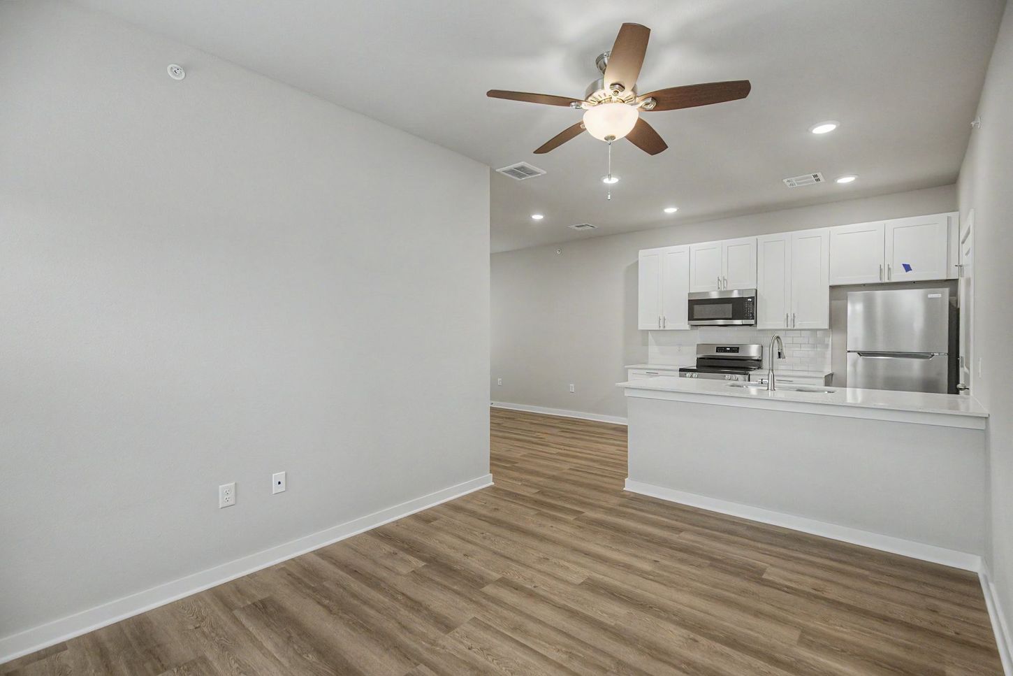 Empty modern living room with wood floors, white walls, ceiling fan, and open kitchen with stainless appliances