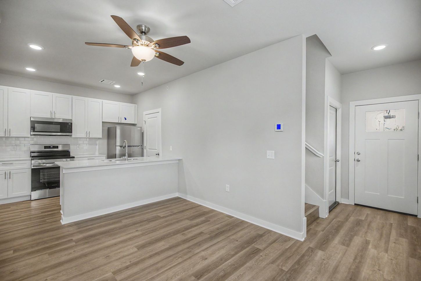 Bright open kitchen and living area with white cabinets, wood flooring, ceiling fan, and front entry door.