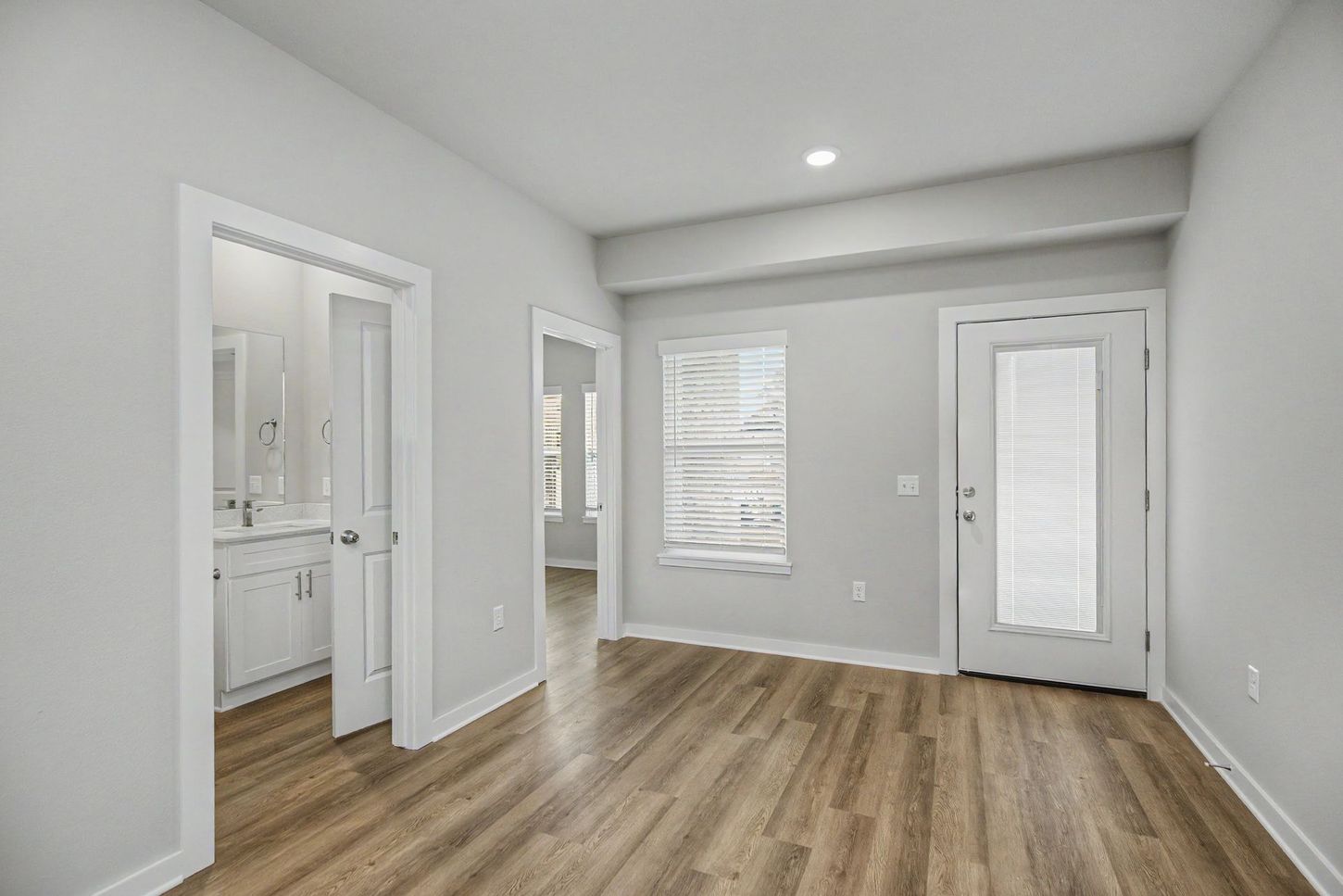 Empty modern room with wood floors, white walls, and a bathroom doorway on the left.