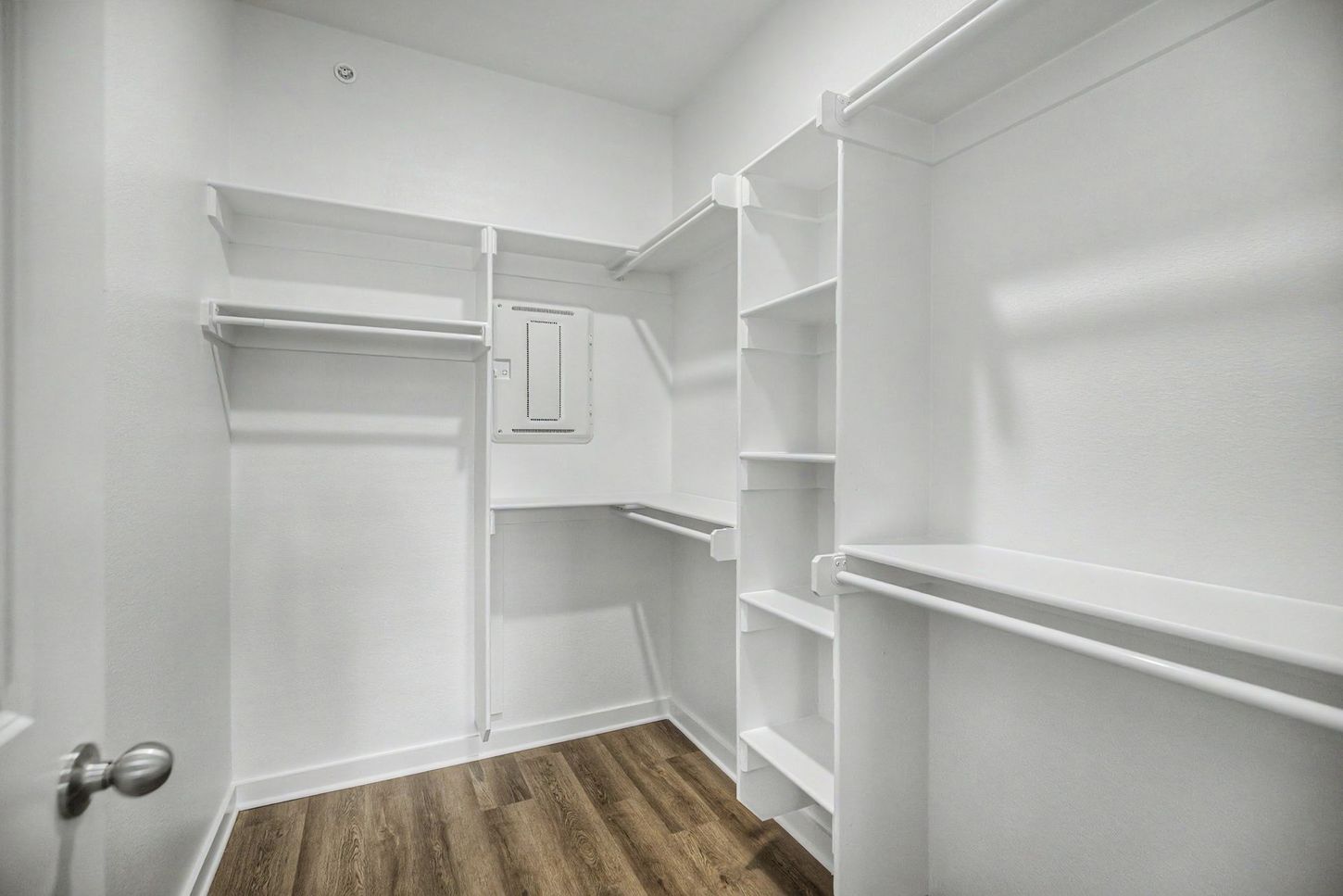 Empty walk-in closet with white shelves, hanging rods, and wood-look flooring.