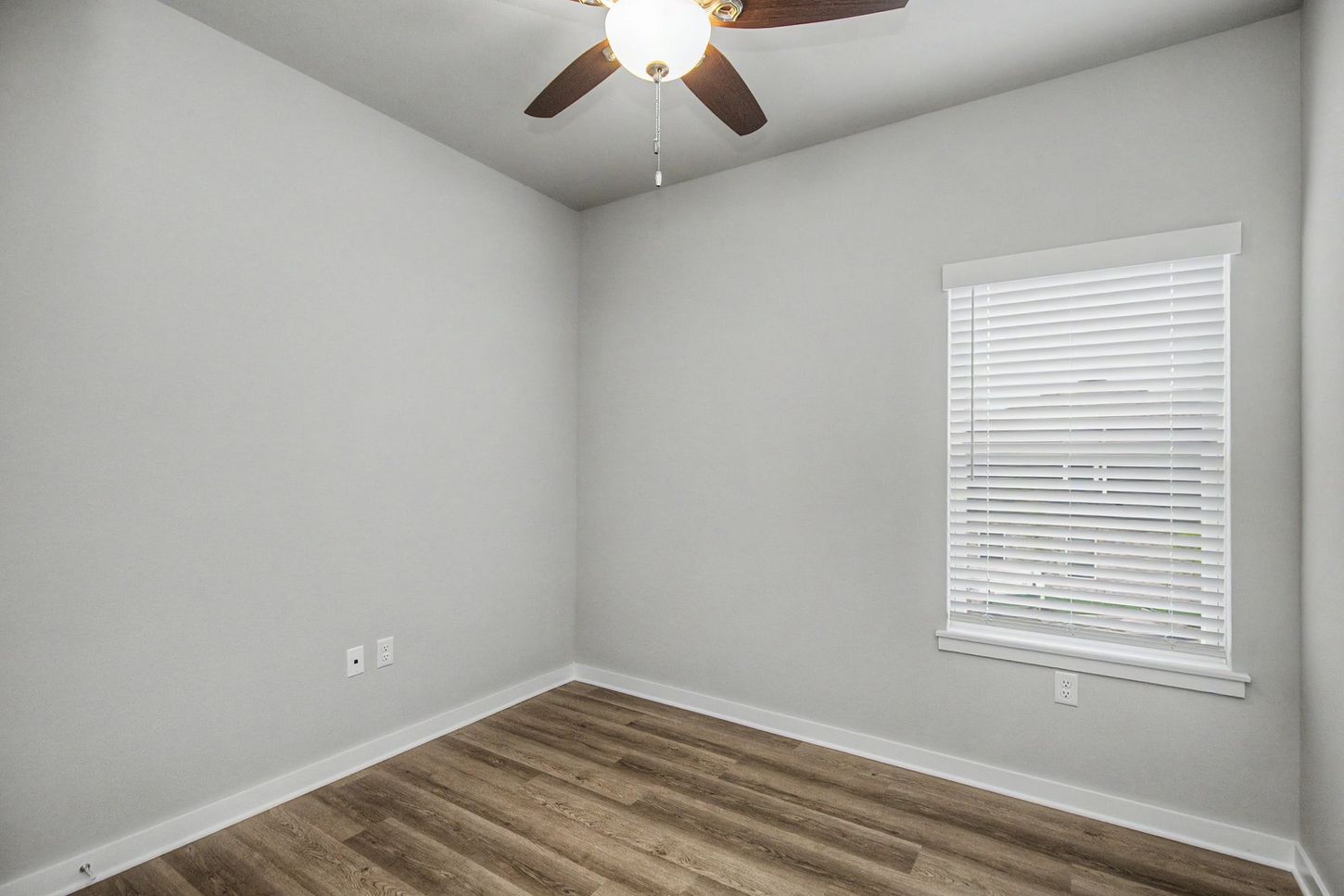 Empty gray bedroom with wood flooring, ceiling fan, and a window with blinds.