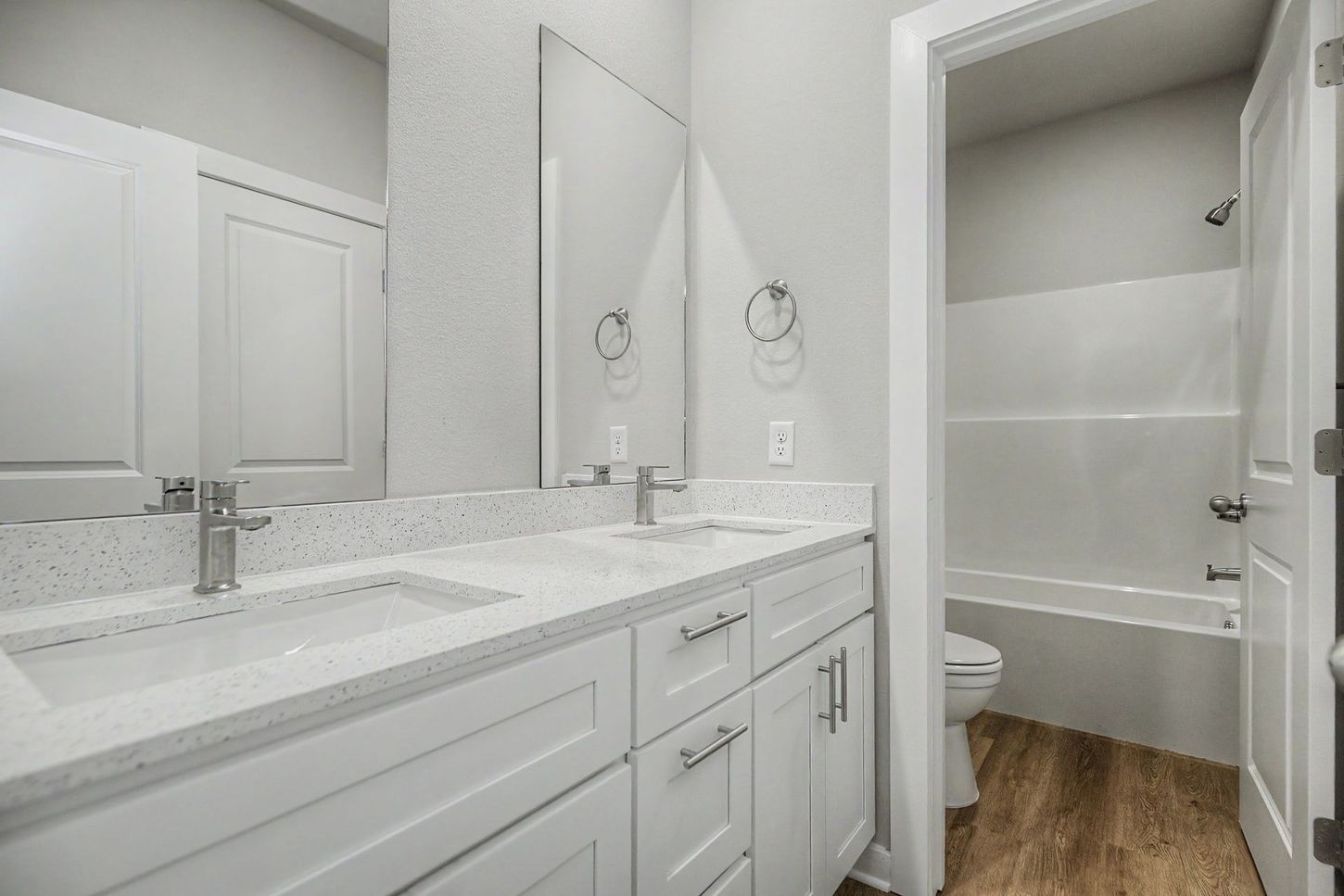 Modern white bathroom with double vanity, large mirror, and shower-tub combo with wood-look floor.