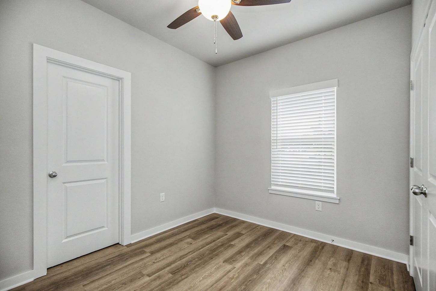 Empty gray bedroom with wood floor, white doors, ceiling fan, and a window with blinds