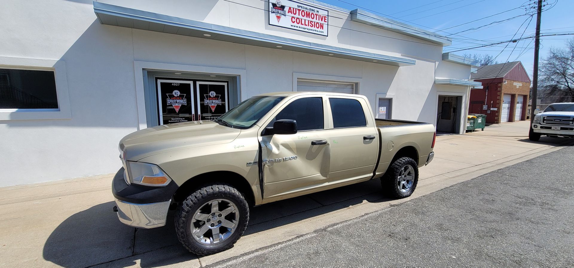 Beige pickup truck with a dented door parked outside a white storefront on a sunny street.