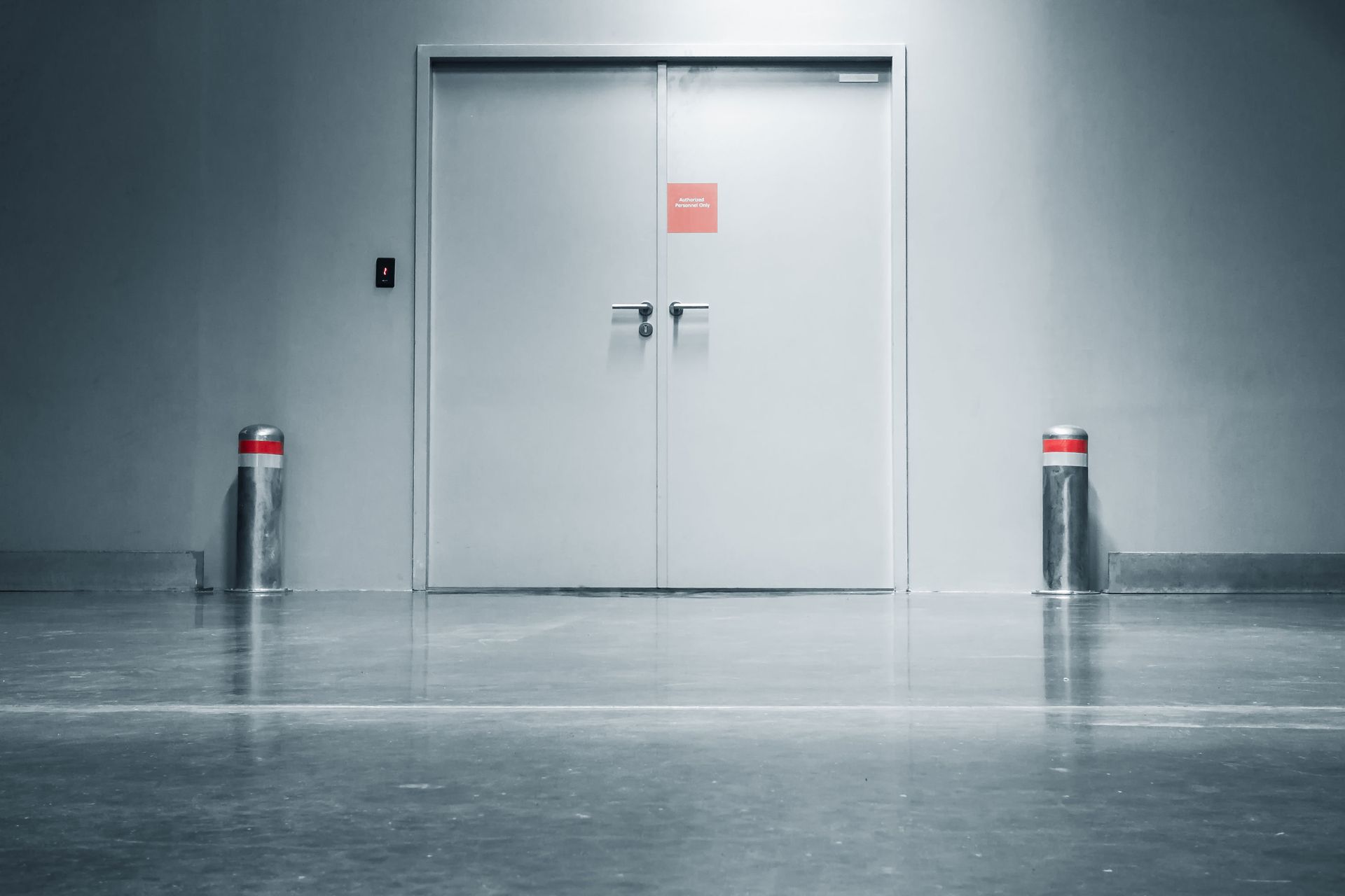 Double doors with a red square, flanked by red-topped bollards, in a gray-walled space. Double doors with a red square, flanked by red-topped bollards, in a gray-walled space.