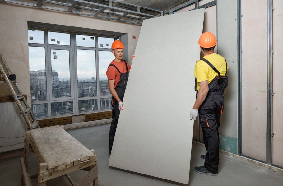 Two construction workers in hard hats carry a large drywall panel inside an unfinished room.