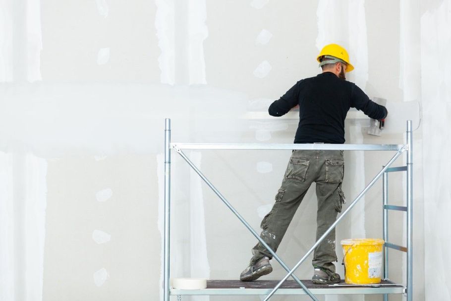 Worker on scaffolding painting a white wall, wearing a yellow hard hat and gray work clothes.
