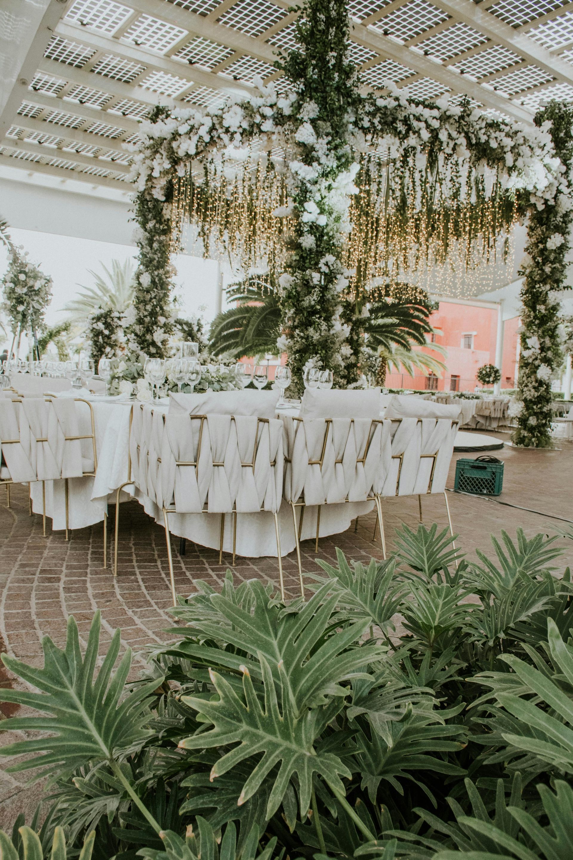 Outdoor wedding reception with white-draped chairs, greenery, and a floral canopy under a pergola