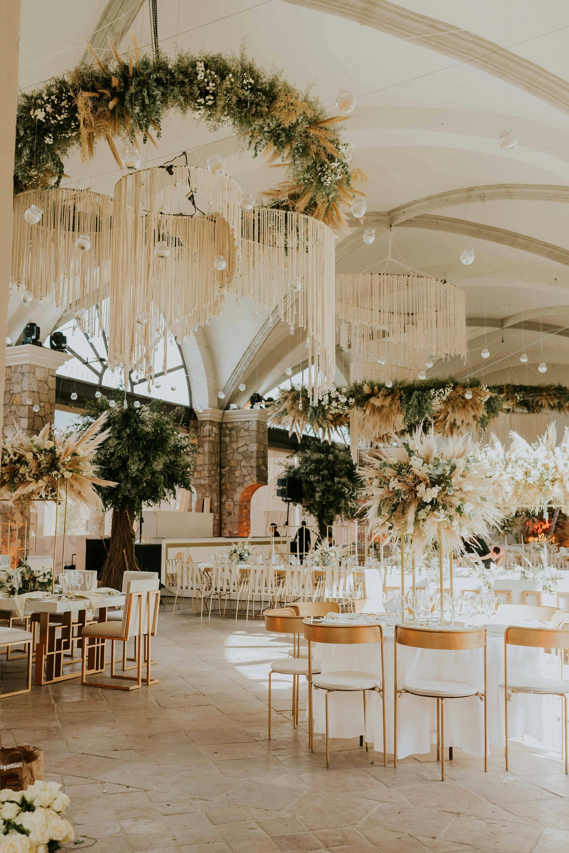Elegant banquet hall with white tables, gold chairs, hanging greenery, and a high arched ceiling