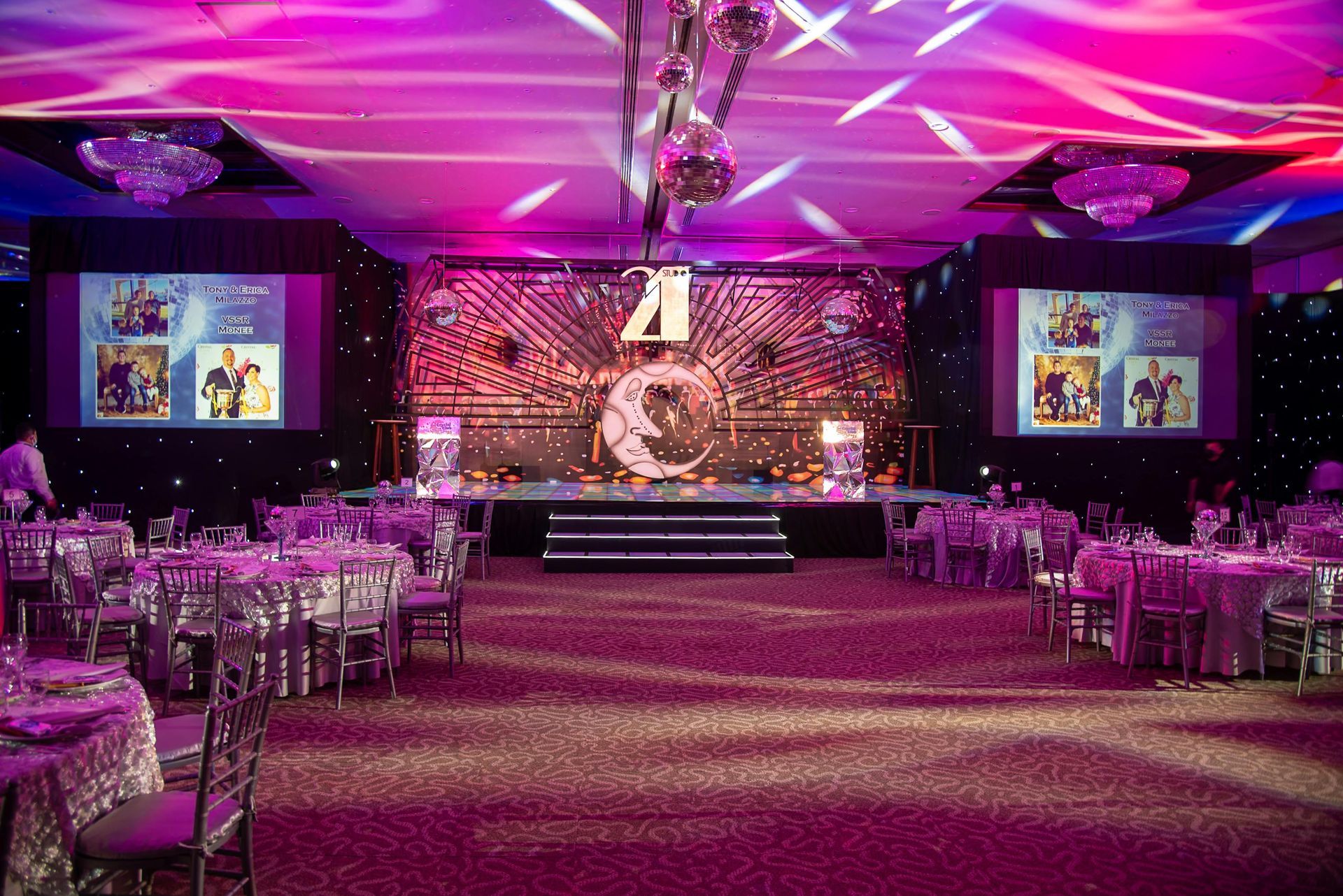 Purple-lit banquet hall with round tables, a stage, and large screens at the front