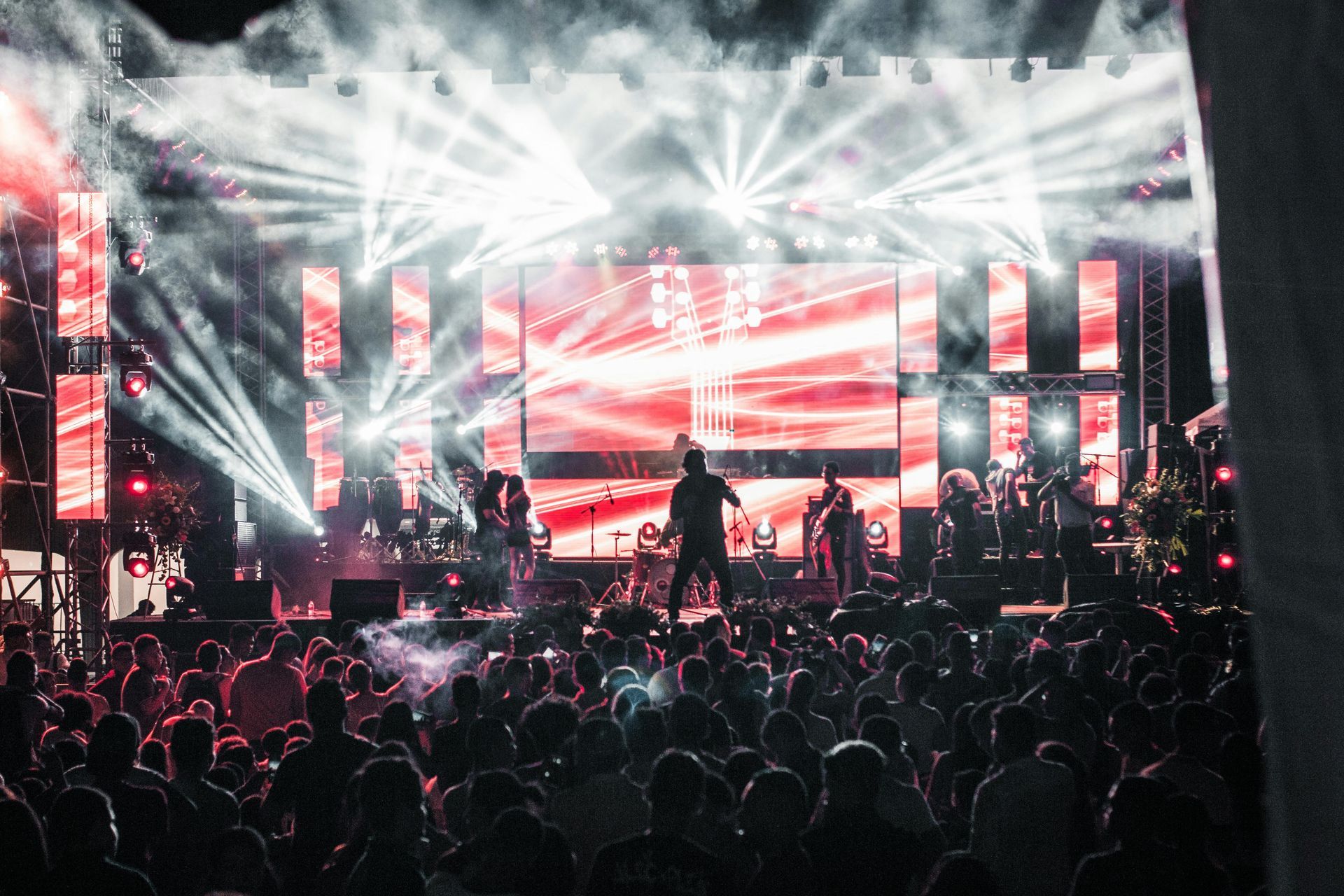 Concert stage with bright white and red lights, performer silhouettes, and a crowded audience