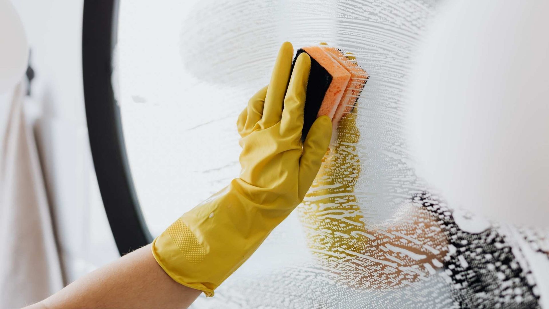 A person wearing a yellow glove uses a sponge to wipe a soapy glass surface.