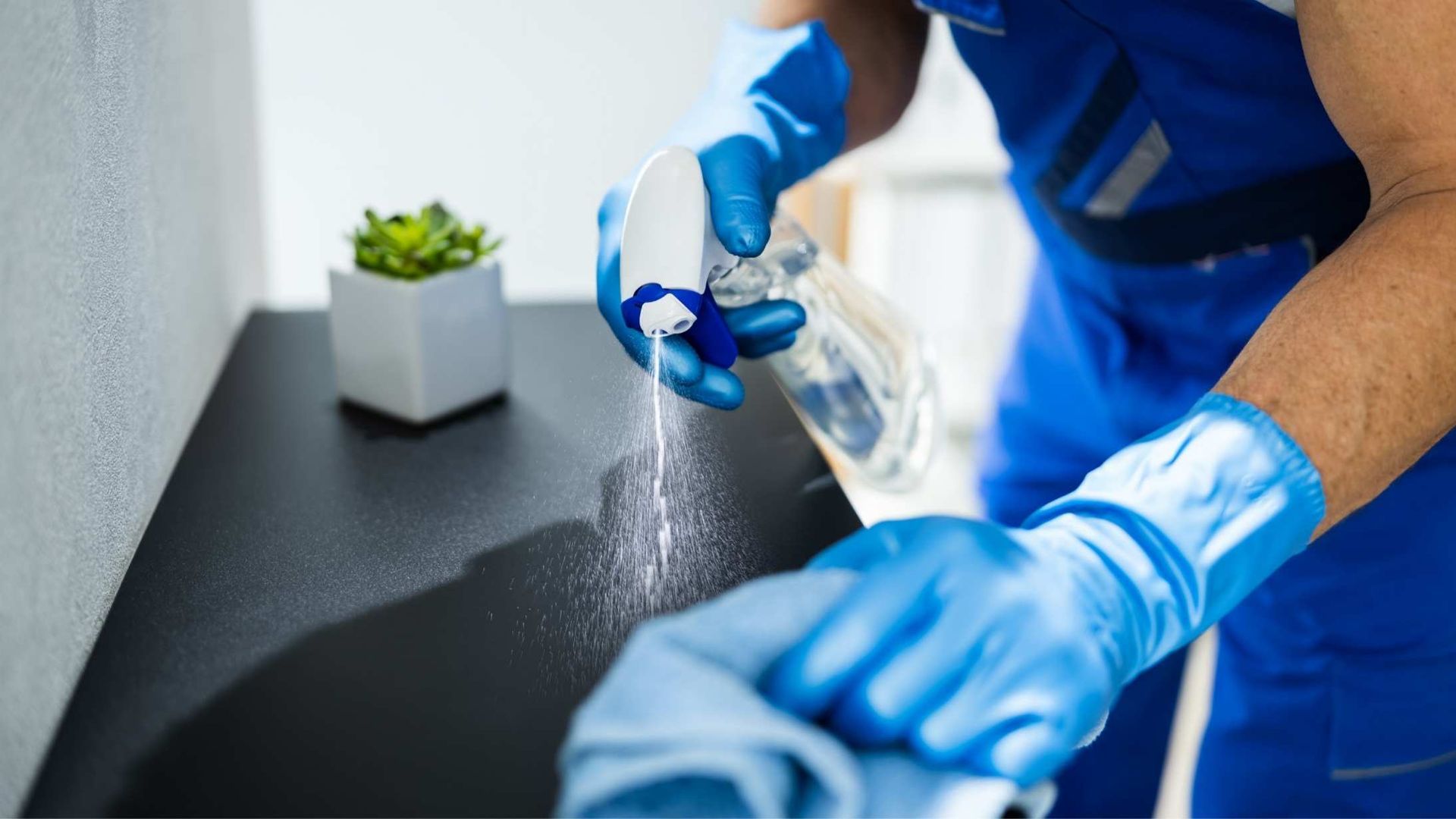 Person wearing blue gloves sprays and wipes a dark surface with cleaning solution.