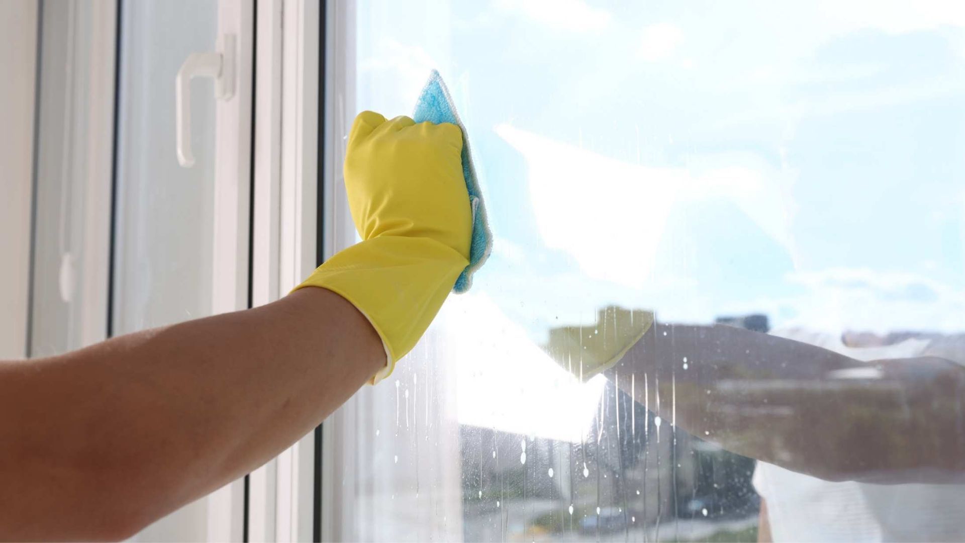 Hand in yellow glove cleaning a window with a blue sponge, bright sunlight reflecting.