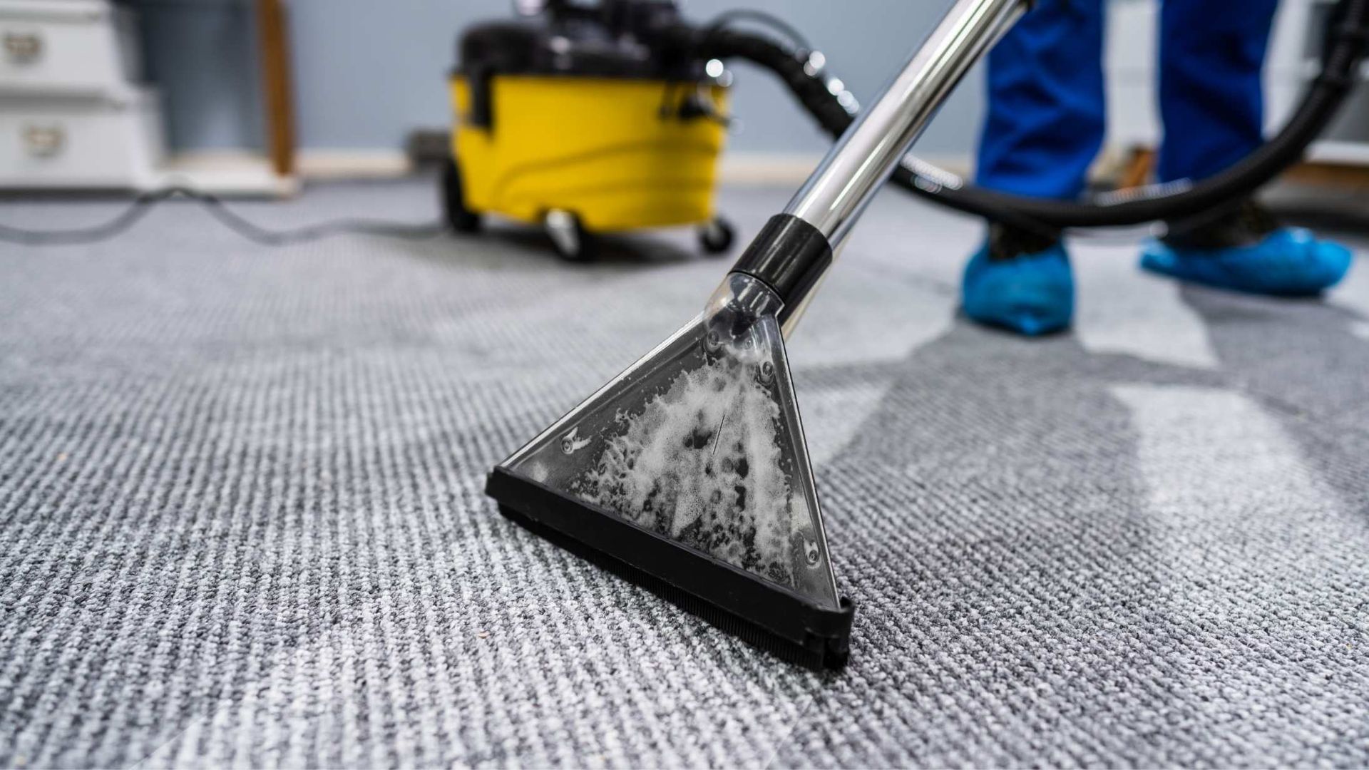 Person using a carpet cleaner on a gray carpet. A yellow vacuum is in the background.