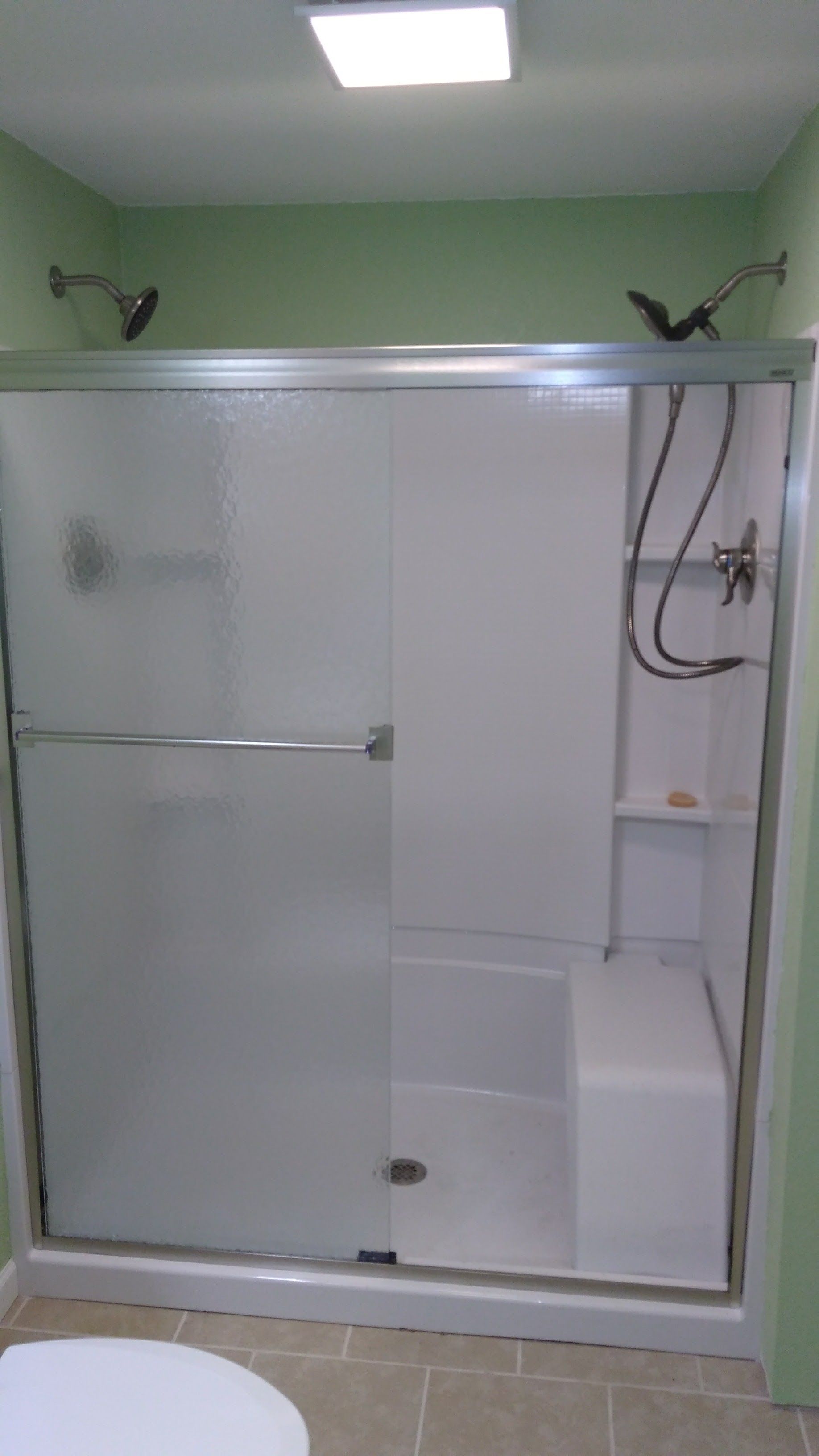 A walk-in shower with a white seat, frosted sliding glass doors, dual shower heads, and green walls.