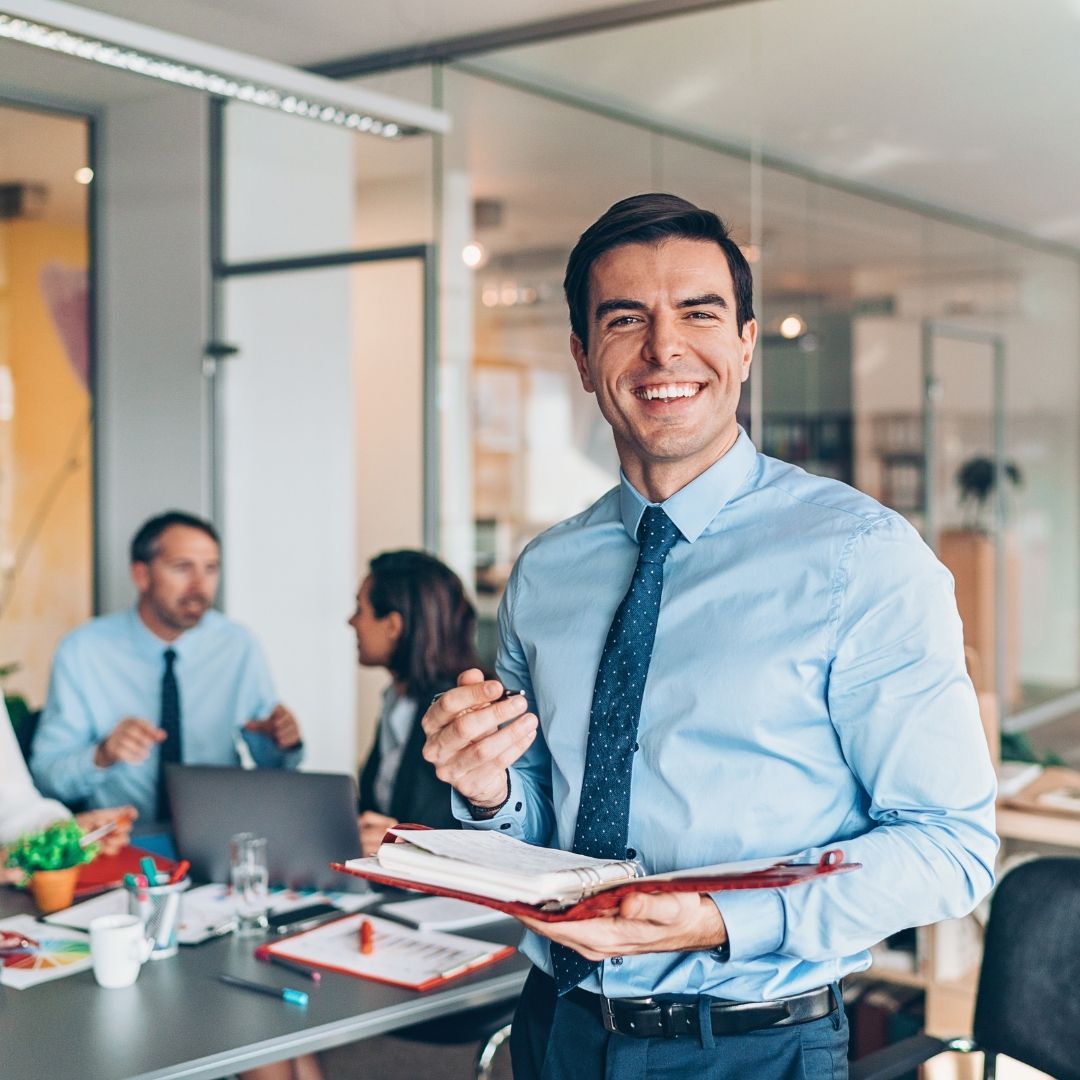 Empresário sorridente segurando um caderno no escritório com equipe trabalhando ao fundo.