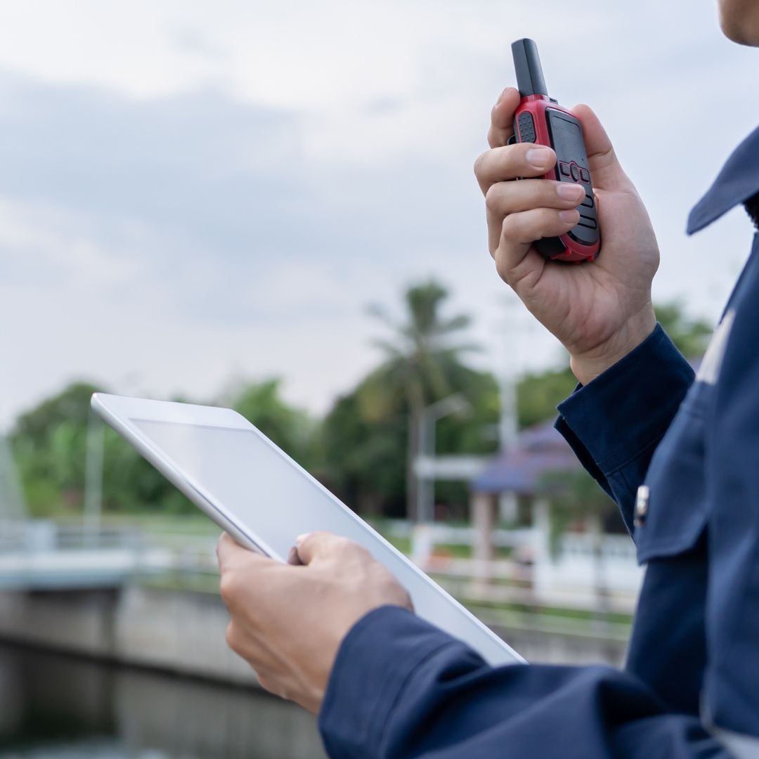 Pessoa de uniforme azul segurando um tablet e um walkie-talkie ao ar livre, perto de uma estrutura - manutenção predial.