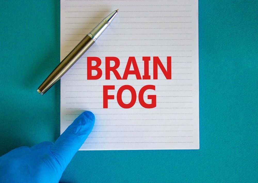 A person is pointing at a notepad with the words brain fog written on it.