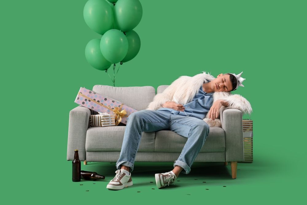 A man is sleeping on a couch with balloons and a bottle of beer.