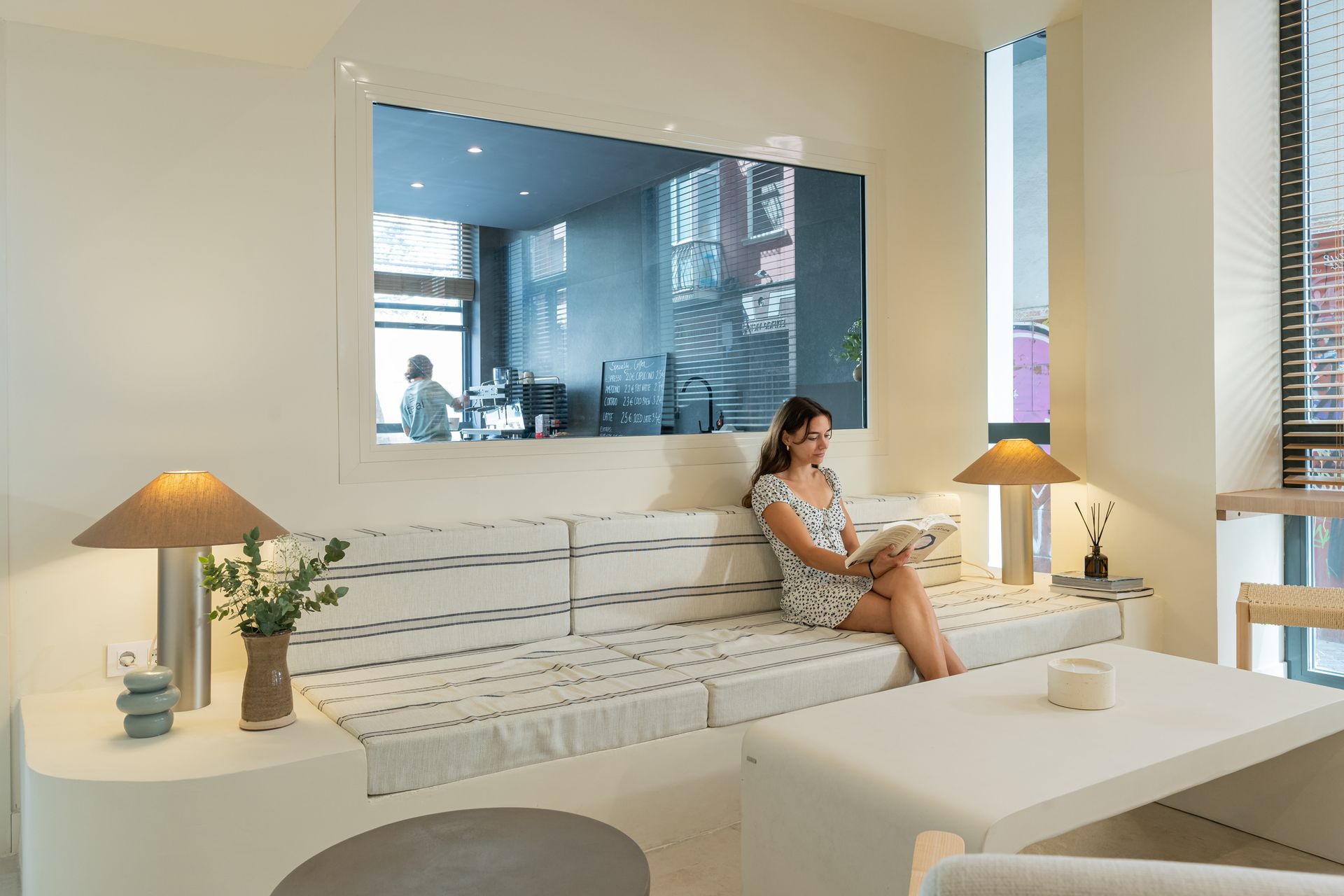 Sala de estar moderna con sofá blanco, lámparas cálidas y una persona sentada en el sofá frente a un gran espejo.