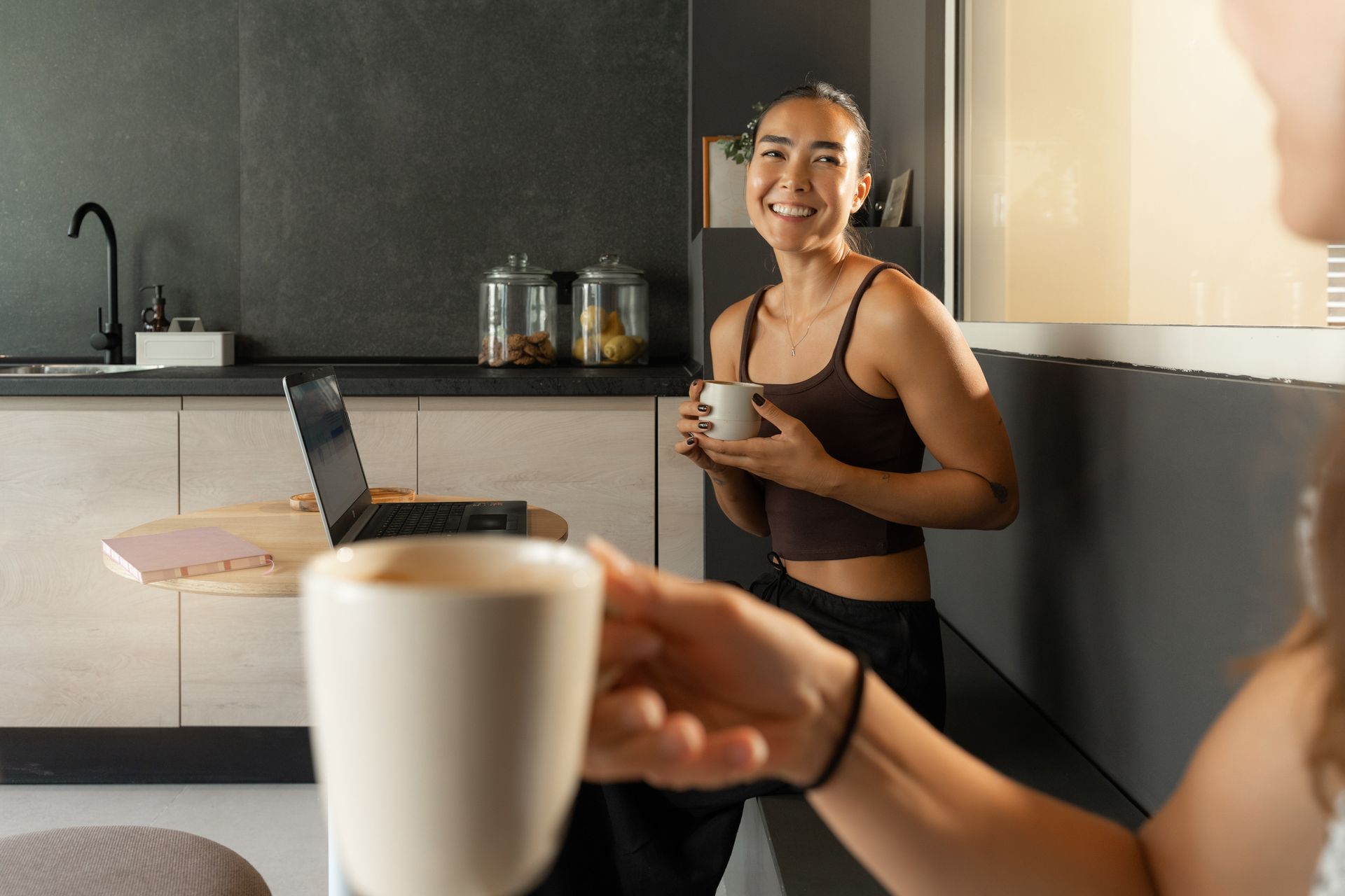 Una persona sonríe en una cocina, sosteniendo una taza mientras otra taza se ofrece en primer plano.