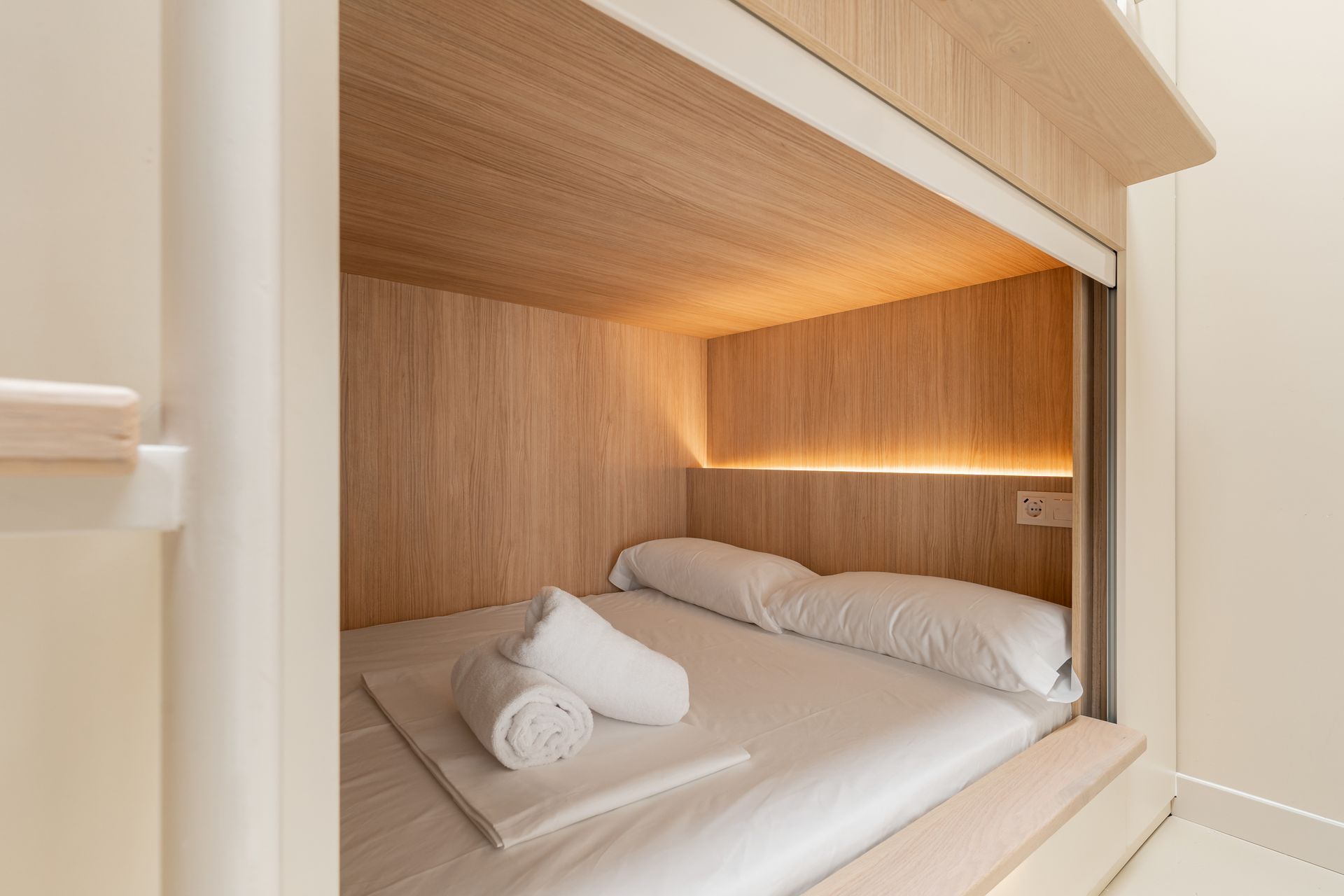 Acogedor rincón con cama integrada, ropa de cama blanca, paneles de madera y cálida iluminación ambiental.