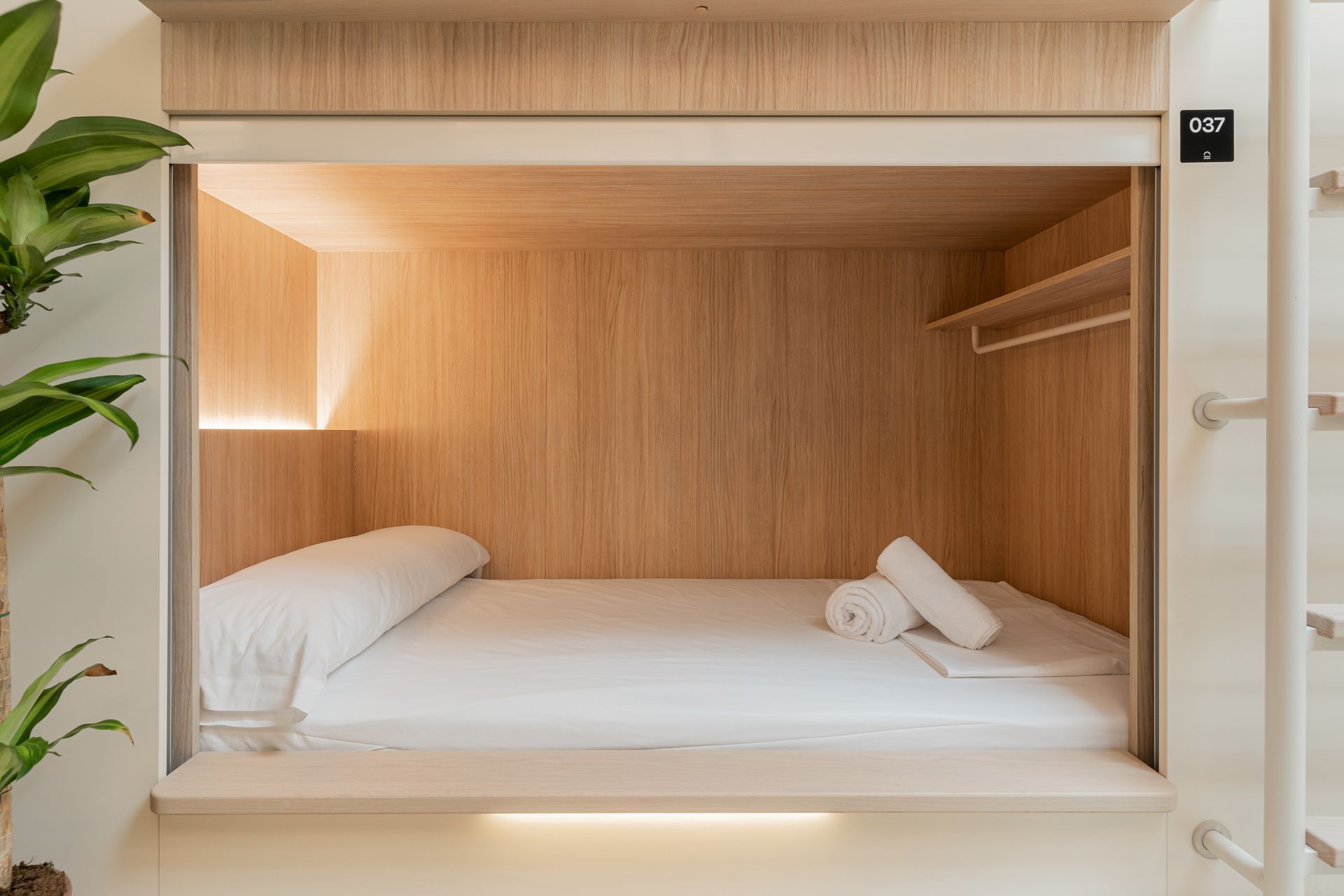 Acogedora cama tipo cápsula de madera con sábanas blancas, almohada, toallas enrolladas y una lámpara colgante dentro de una alcoba abierta integrada.