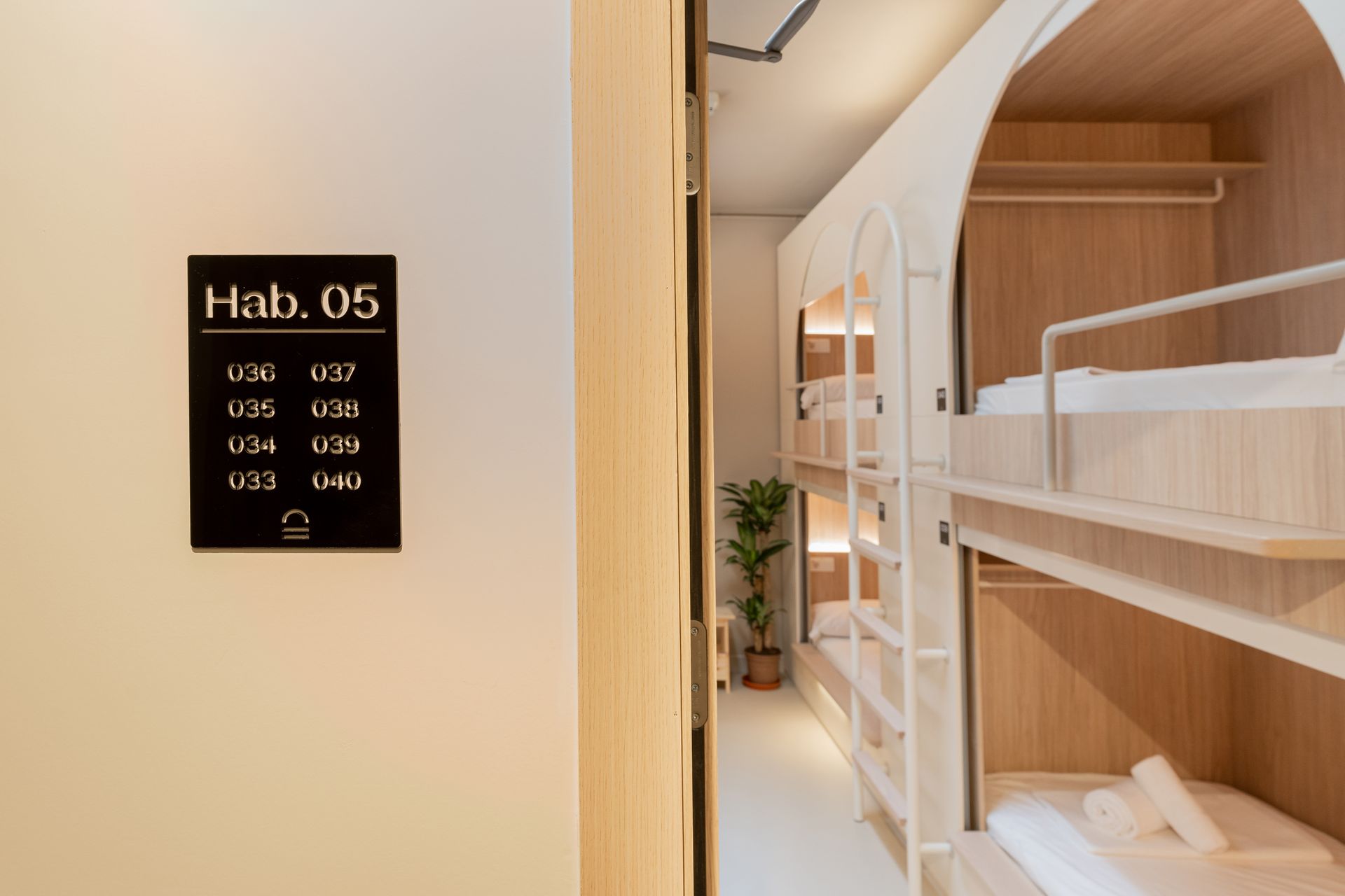 Letrero de Hab. 05 junto a una luminosa habitación de dormitorio de madera con literas y sábanas blancas.
