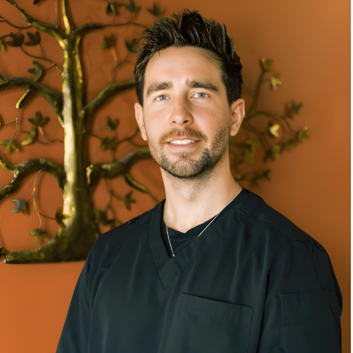 Man in black top, smiling, stands in front of a wall with golden tree decoration.