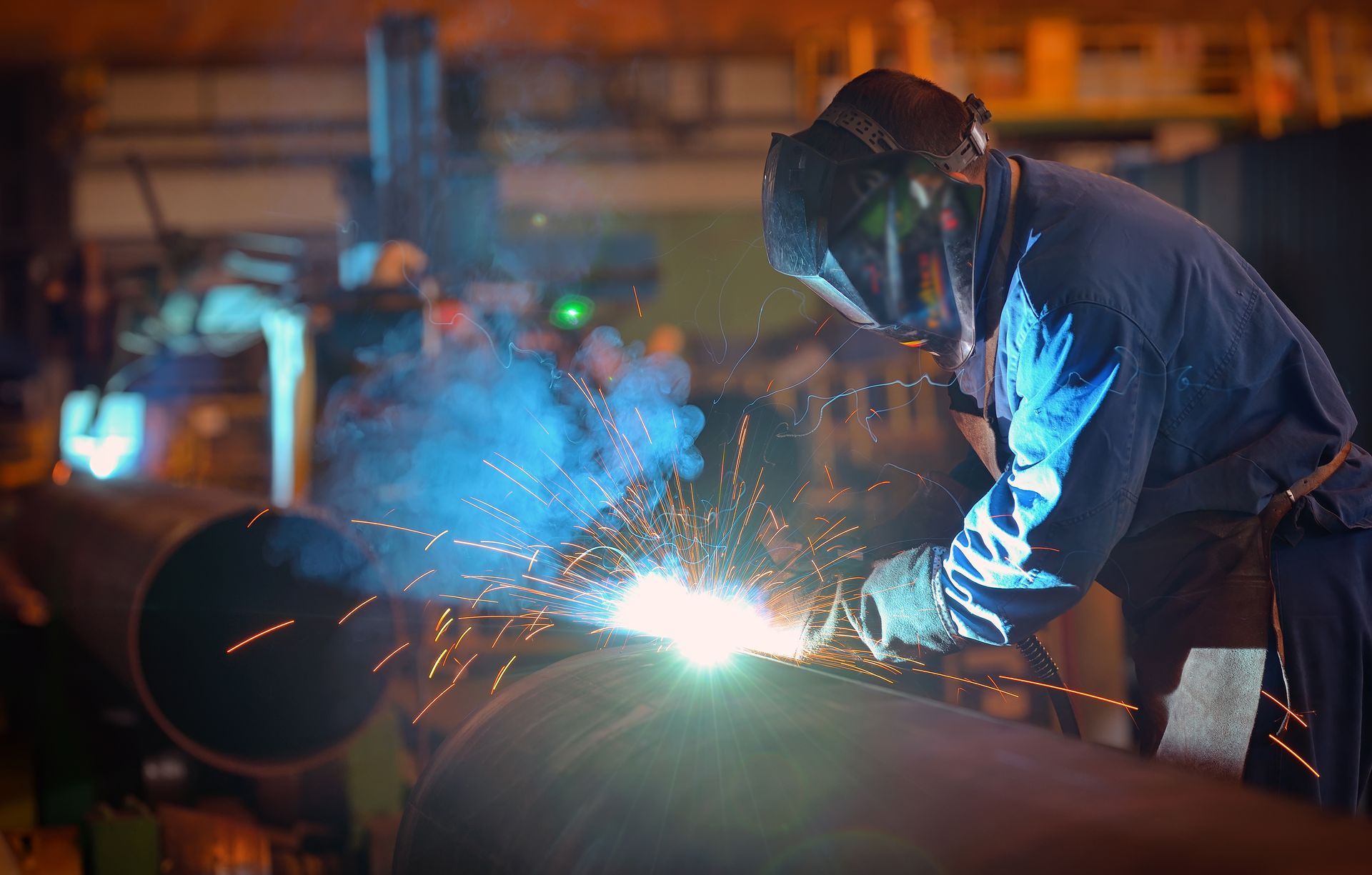 Welder in a protective hood sparks bright blue light while working on a large metal pipe in a workshop