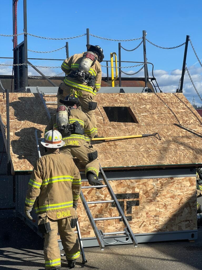 Firefighters on a roof using a ladder during training, with one climbing and one standing below.