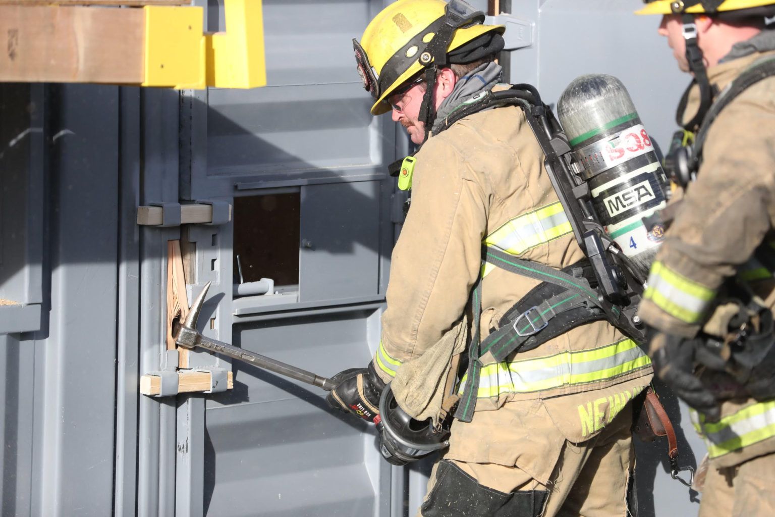 Two firefighters in tan gear use a tool to pry open a gray metal door