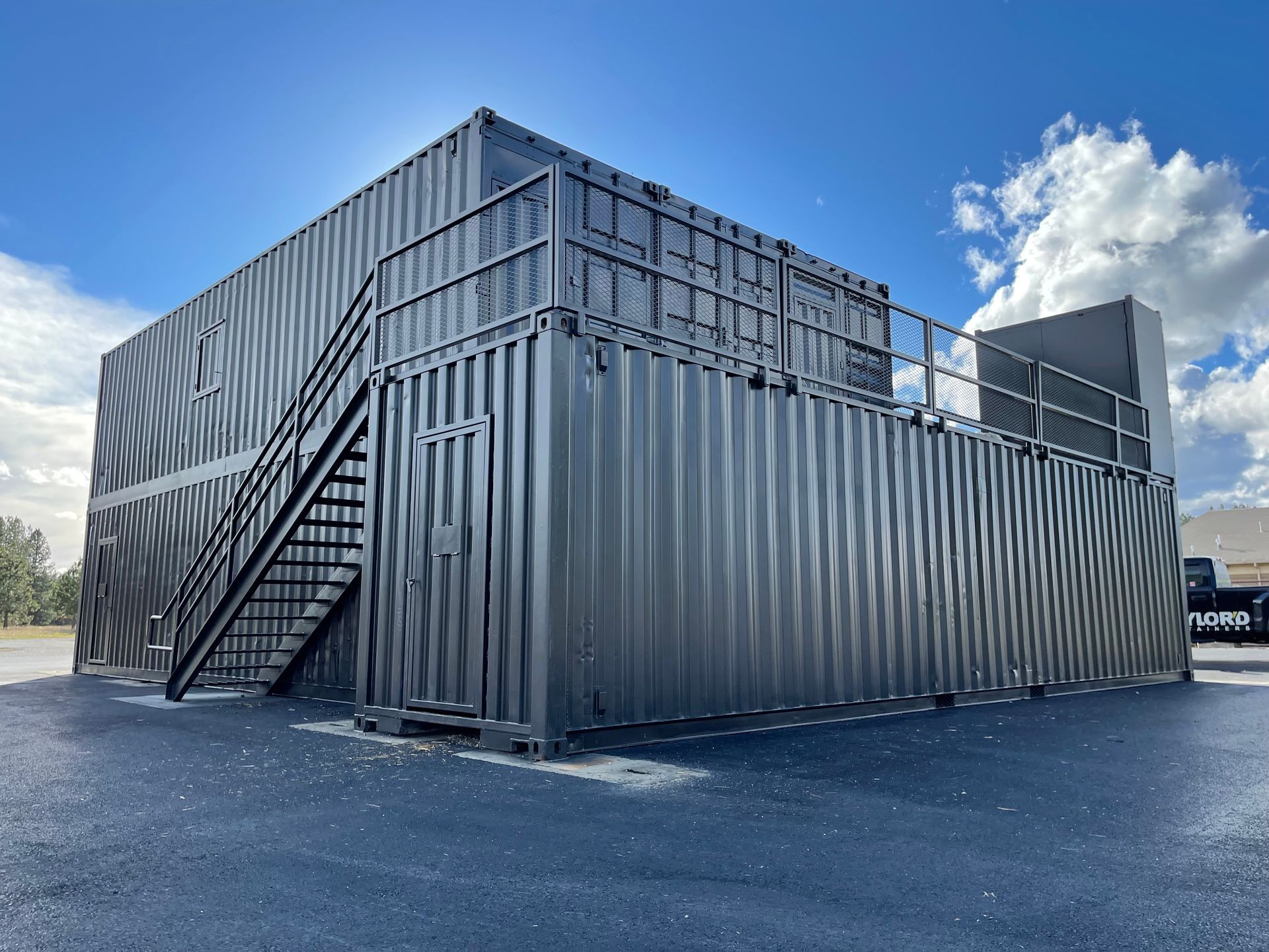 Stacked gray shipping containers with exterior stairs, set on a paved lot under a blue sky.