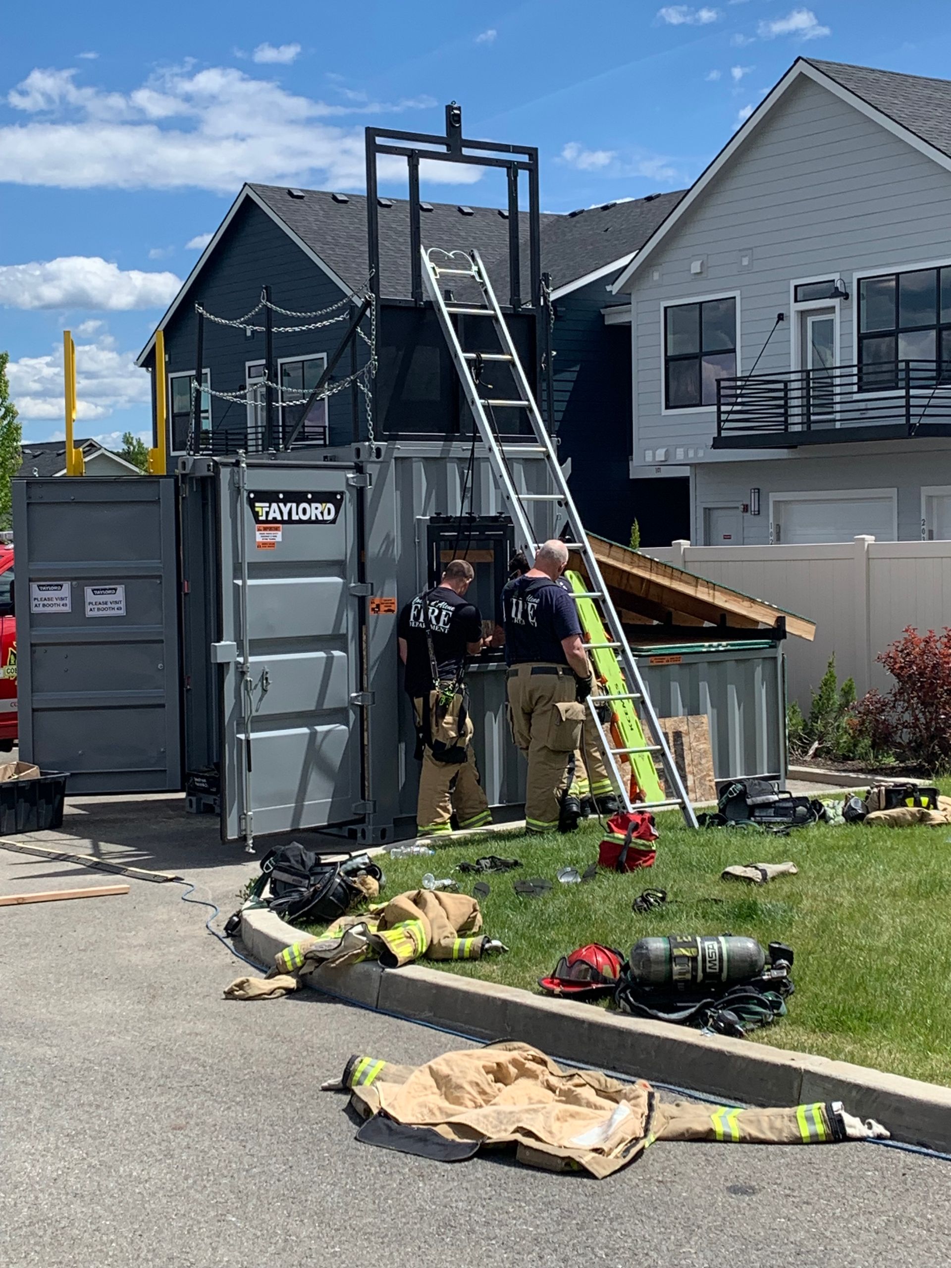 Workers using a ladder beside a dumpster outside a townhouse under construction, with tools on the ground