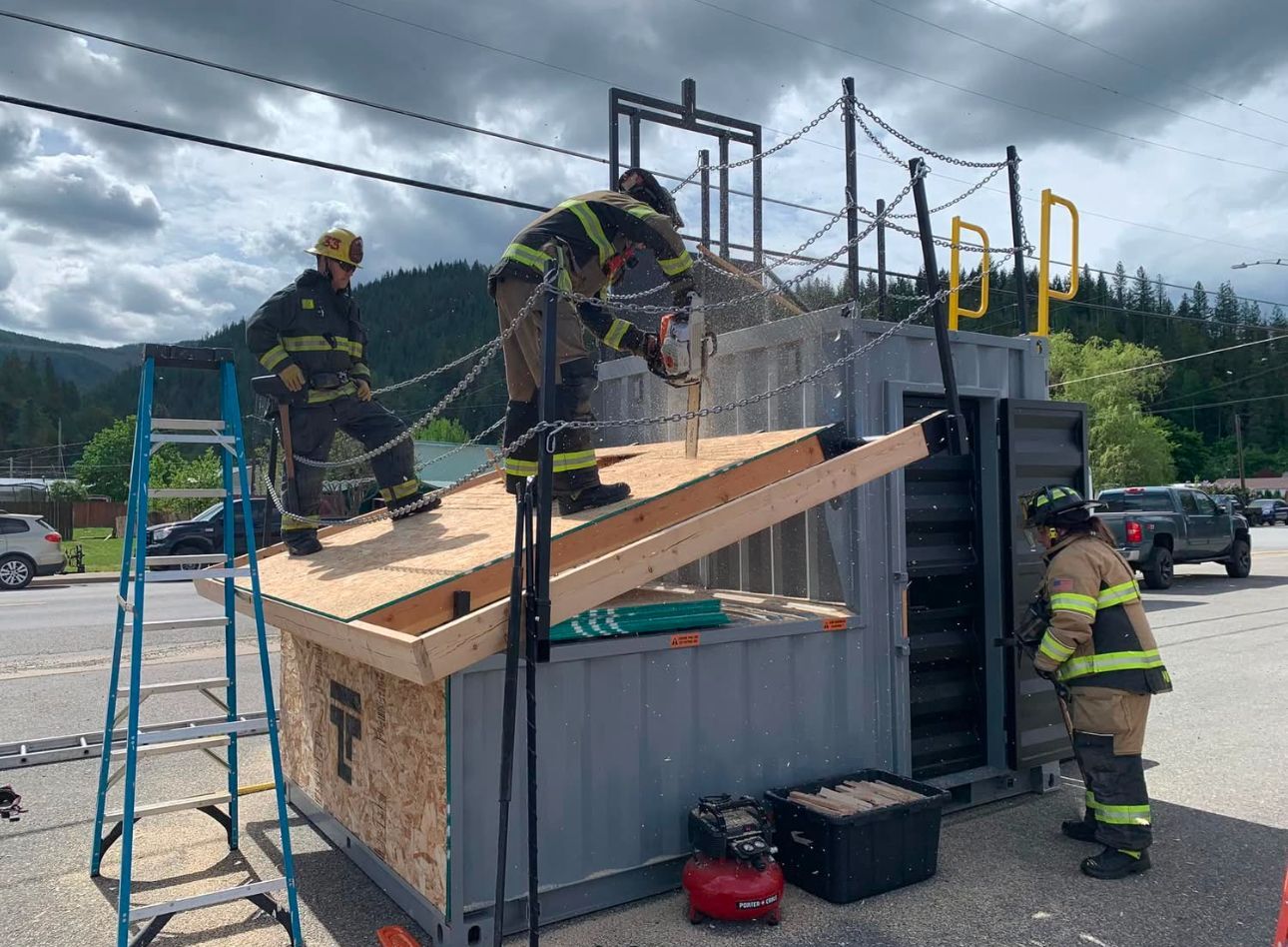 Firefighters on a tilted overturned truck, inspecting the cab in a parking lot under cloudy skies.