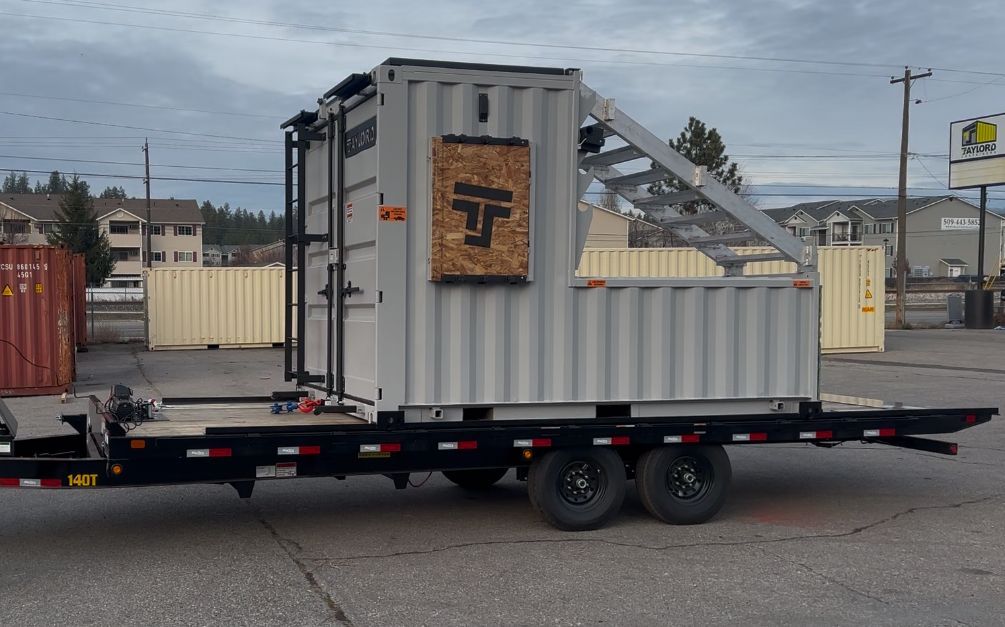 Flatbed trailer carrying a small gray shed-like structure in a parking lot under a cloudy sky