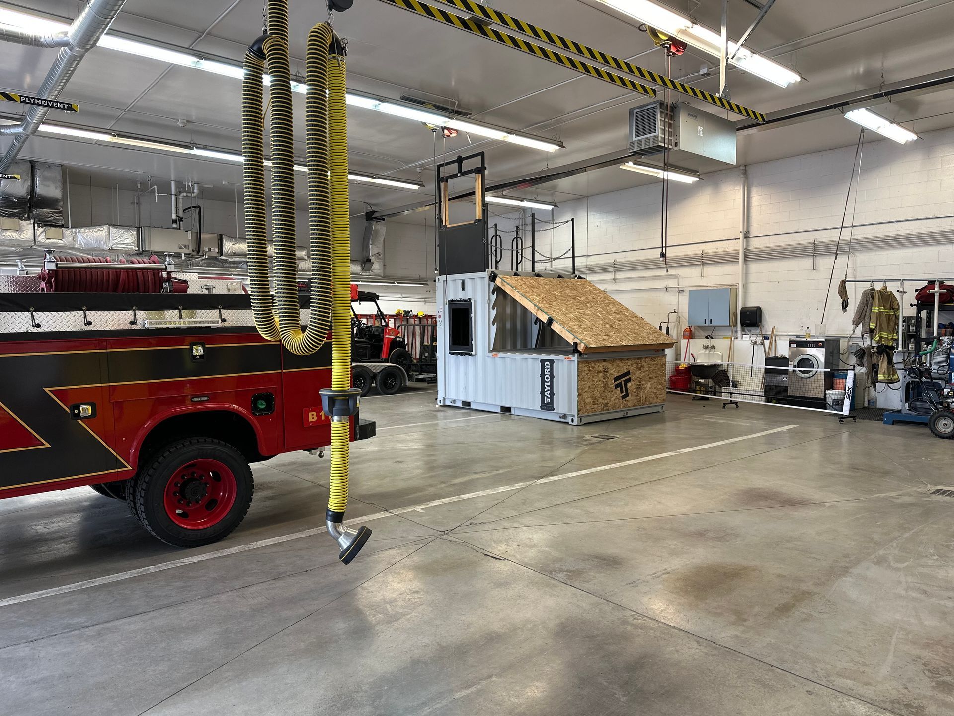 Fire station garage with a red fire truck, hanging hoses, and a training structure inside.