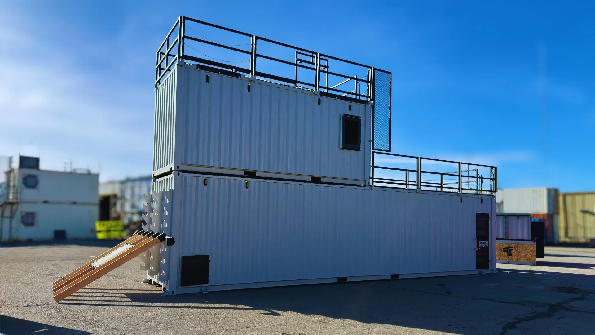 White modular building with rooftop railings and exterior stairs in an industrial yard under a blue sky