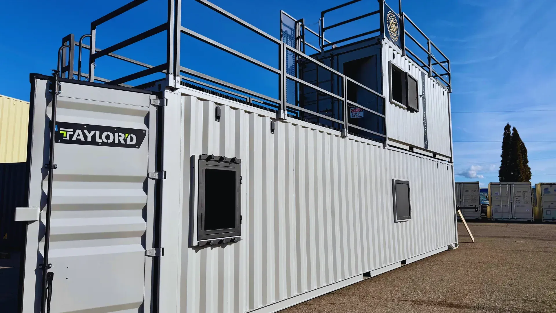 White shipping container office with blue railings and a Taylor logo, set outdoors on gravel under a clear sky